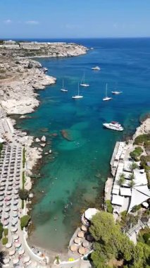 Güneşli bir günde, Rodos 'taki Kallithea Sahili' nin turkuaz su, kayalık kıyı şeridi, yatlar, şemsiyeler ve yaz tatili atmosferiyle insansız hava aracı görüntüsü.