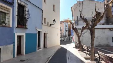 Geleneksel İspanyol sahil kasabasında renkli evleri ve bankları olan sessiz dar bir cadde. Güneşli Akdeniz mimarisi ve huzurlu şehir atmosferi.
