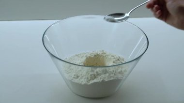El, beyaz mutfak arka planında hamur hazırlığı sırasında cam kaseye kaşıkla tuz ekliyor. Pişirme işlemi kapat.
