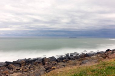Uzak bir kargo gemisi dramatik bulutlu bir gökyüzünün altında sakin bir denizde yüzüyor. Ön planda uzun süre maruz kalınca yumuşayan kıyı kayaları, sakin ve ruhani bir kıyı şeridi sahnesi oluşturuyor..