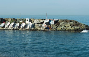 6 Aralık 2025, Trabzon, Türkiye. İnsan yapımı taştan bir dalgakıranın ya da sakin denize uzanan bir iskelenin yakın plan görüntüsü. Birçoğu beyaz muşambalarla kaplı küçük balıkçı tekneleri, derme çatma ahşap bir yapının yanında iç kısımda korunuyor.