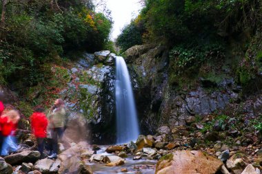 Kayalık bir orman geçidinde akan bir şelalenin sakin ve uzun pozlu bir fotoğrafı. Önplanda hafif hareket bulanıklığında renkli giysiler içinde yürüyüş yapan yürüyüşçüler var. İpeksi su ve dinamik insan hareketi arasındaki zıtlık...