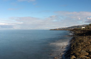 Karadeniz 'in engebeli, kayalık kıyı şeridinin uzun pozlu fotoğrafı denizin bulanık hareketini gösteriyor. Yoğun bir yerleşim yeri mavi gökyüzünün altındaki yükselen tepeleri çiziyor..