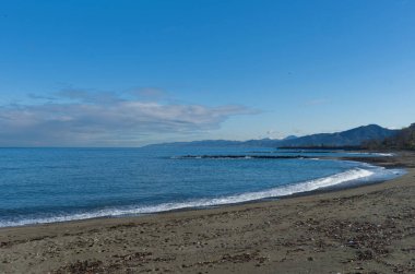 Expansive horizontal shot of a dark sandy beach on the coastline, with gentle waves breaking near the shore. The clear horizon meets a backdrop of rolling hills and mountains under a blue sky.