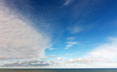 Dramatik mavi gökyüzünün geniş, yüksek kontrastlı panoramik görüntüsü, soldaki koyu, yoğun bulutlardan sağdaki açık mavi gökyüzüne, durgun deniz ufkunun üzerinde keskin bir geçiş içeriyor..