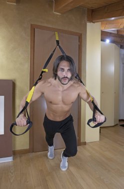 Genç atletik adam evde süspansiyon kayışları ile antrenman yapıyor, üstsüz fitness modeli TRX bantlarıyla göğüs şınavı yapıyor, kapalı alanda egzersiz yapıyor, sağlıklı yaşam tarzı, kas ve vücut eğitimi alıyor..