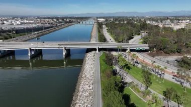 Long Beach, California 'daki Los Angeles Nehri bisiklet yolunun 710 no' lu otoban boyunca uzanan insansız hava aracı görüntüsü. Bisikletçiler kaldırımlı yolda giderken, arabalar otoyolda seyahat ediyor, SoCal 'da kentsel ulaşım ve açık hava eğlencesi vurgulanıyor
