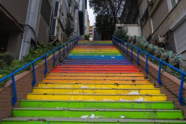 Gökkuşağı desenli, binalar ve yeşilliklerle çevrili renkli açık hava merdivenleri. Mavi korkuluklu renkli gökkuşağı merdivenleri