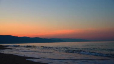 Huzurlu deniz dalgaları, karanlık kumsal kumları ve ufukta katmanlı dağlarla kıyı günbatımı sahnesi. Güzel altın saat deniz manzarası ve manzarası