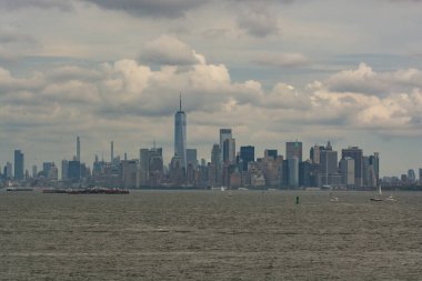 New York şehrinin geniş bir manzarası bulutlu bir gökyüzü altında, ikonik gökdelenler ve huzurlu bir deniz manzarası.