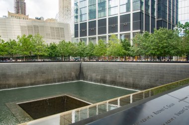 New York 'ta yeşil ağaçlar ve canlı kentsel mimari ile çevrili 11 Eylül Anıt Pınarı' nın sakin manzarası, yansıma ve hatırlama hissini uyandırıyor..
