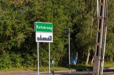Yeşil bir işaret Kotobrzeg kasabasını gösteriyor, gür ağaçların arasında uzun süre ayakta duruyor. Huzurlu çevreyi yansıtır ve yolculara yön gösterir..