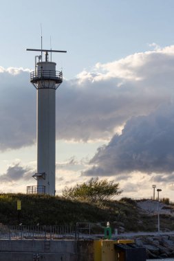 Limanın yakınında çimenler ve kayalarla çevrili bir deniz feneri var. Gökyüzünde öğleden sonra saatlerinde dramatik bulutlar beliriyor, sakin bir kıyı manzarası yaratıyor..