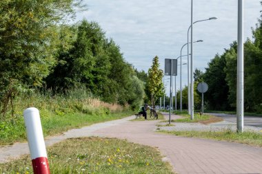 Yeşillik ve ağaçlarla çevrili bisiklet yolu boyunca bir bankta oturan bir insan. Gökyüzü açık ve yakınlarda bir bisiklet park edilmiş. Bugün sakin bir gün..