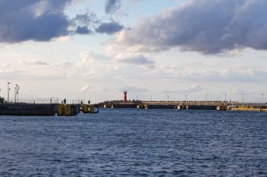 Hareketli bir liman sahnesi, teknelerin kenetlendiğini ve arka planda bulutlu bir gökyüzünün altında bir deniz feneri olduğunu gösteriyor. İnsanlar öğleden sonra sıcacık atmosferin tadını çıkararak rıhtımda sıraya giriyor..