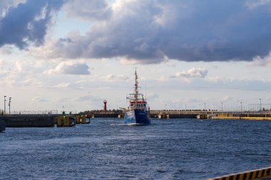 Bir sahil gemisi gece yaklaşırken dikkatlice limana doğru ilerliyor. Gökyüzü bulutlarla dolu ve kıyı şeridinden uzak bir kalabalık izliyor..