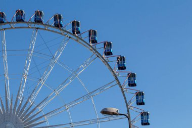 Parlak mavi gondolları olan dev bir dönme dolap açık mavi bir gökyüzünün altında belirgin bir şekilde duruyor. Güneşli bir gün, eğlence parkında gezinti yapmak için mükemmel..