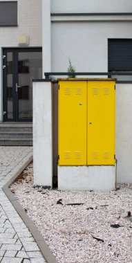 Modern bir binanın yanında parlak sarı bir hizmet kutusu bulunmaktadır. Çevresindeki alan beyaz çakıl ve parlak, minimal mimariye sahip ve sarı ile bir kontrast oluşturuyor..