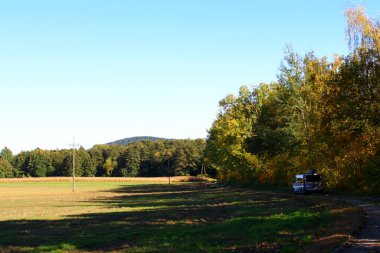 Sakin bir kırsal bölgede güneşli bir tarla boyunca canlı sonbahar ağaçları yer alır. Park halindeki bir araç, ağaç sınırının yanında görülebiliyor. Sessiz sedasız güzellikler sergiliyor..