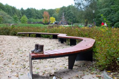 Bir çift sağlam bot sessiz bir parktaki eğri bir ahşap bankta oturuyor. Sonbahar yaprakları çevreyi dolduruyor ve ağaçlar renkli yapraklar sergiliyor. İnsanlar uzaktaki parkın tadını çıkarıyorlar..