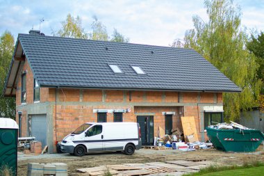 İnşaat alanında inşa sürecinde iki katlı bir ev yer alıyor. Yakınlarda beyaz bir minibüs var ve çöp konteynırında inşaat atıkları var. Arka planda ağaçlar var.