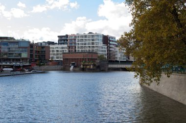 Gökyüzünü ve ağaçları yansıtan sakin bir nehrin yanında binalar yükselir. Bölge, açık bir öğleden sonra doğa ve şehir hayatının bir karışımını gösteriyor. Huzurlu bir manzara yaratıyor..