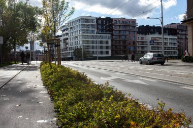 İki yaya, kentteki sessiz bir yolun yanında düzenli bir patika boyunca yürür. Modern binalar arka planda yükselir ve ağaçlar caddede sıraya girer. Bulutlarla kaplı sakin bir gün..