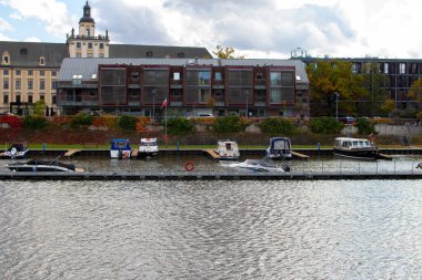 Arka planda modern binalar ve ağaçlar bulunan sakin nehir boyunca bir sıra tekne kenetlendi. Bazı bulutlarla açık bir gün, huzurlu bir atmosfer yaratıyor..