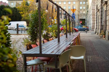 Long wooden table with colorful chairs is set up along a peaceful city street. Trees display autumn colors, creating a cozy atmosphere perfect for outdoor dining.