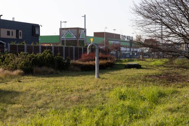 An open field surrounds a shopping area, with shrubs and grass contrasting against nearby commercial buildings. Its a sunny day, highlighting the blend of nature and urban life.
