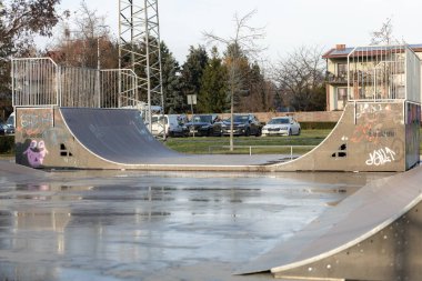 Kavisli rampaların yer aldığı kaykay parkı banliyö bölgesinde bulutlu bir gökyüzünün altında duruyor. Islak zemin yakınlardaki binaları ve ağaçları yansıtıyor, yeni yağmuru gösteriyor..