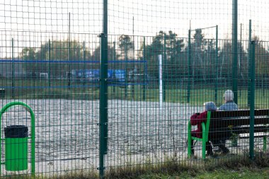 Yaşlı bir çift, öğleden sonra bir voleybol maçını izlemek için bir bankta yan yana oturur. Huzurlu ortam bir topluluk ve rahatlama hissi sunuyor..