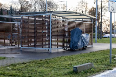 Boş bir otoparkta, mavi bir brandayla kaplı bir bisikletle bir bisiklet sığınağı duruyor. Sahne, ağaçlar ve seyrek araçlarla çevrili günbatımının sükunetini yakalıyor..