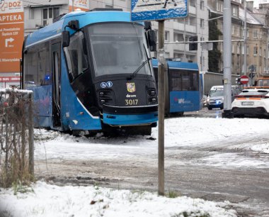 Mavi bir tramvay karlı bir caddede sıkışmış, hareket etmeye çabalıyor. Etrafı karla kaplı olan bölge, Polonya 'nın bir kentindeki kış koşullarını vurguluyor..