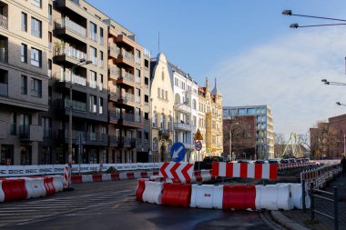 Şehir merkezinde yol inşaatı devam ediyor. Yollara trafik engelleri konuluyor. Yakınlardaki binalar eski ve yeni tasarımların bir karışımını gösteriyor. Çalışmalar devam ederken insanlar geçebilir..