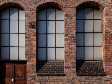 Büyük kemerli pencereleri ve ahşap kapısı olan eski bir tuğla bina. Güneş ışığı tuğla duvarda gölgeler oluşturur. Yapının detaylarını vurgular..