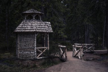Kaba, el yapımı ahşap bir çardak (barınak ya da küçük bir kulübe), küçük ahşap bir köprüyle bağlanmış kiremitli bir çatı ile dramatik ve geniş bir görüntü. Sahne karanlık, yoğun ve ürkütücü kozalaklı bir ormanda geçiyor.