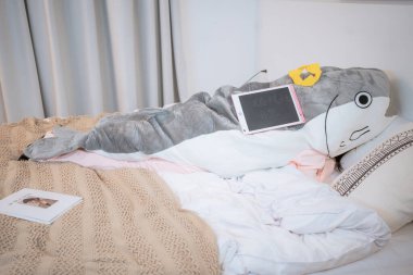 Dev bir köpekbalığı pelüşü ve yatağın üzerinde modern bir tabletin bulunduğu samimi bir sahne rahatlık ve masumiyeti çağrıştırıyor. Yaşam tarzı ve ev dekorasyonu için ideal.