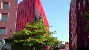 Açık mavi gökyüzüne karşı koyu kırmızı geometrik binalarla modern mimarinin 4K görüntüsü. Temiz çizgiler, pencereler ve eşsiz cantilever tasarım elemanlarıyla minimal kentsel sahne. 