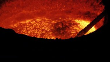 Parlayan turuncu ve sarı erimiş metal bir yüzey ve karanlık bir endüstriyel ortamda içine bir çubuk batırılmış..