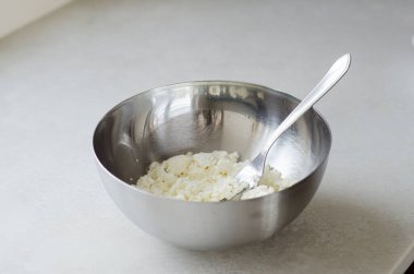  Kremalı, kaba, süzme peynir. Parlak bir mutfak masasında metal bir kasede çatalla ezilmiş. Sağlıklı yemek ve süzme peynir pişirme tarifleri için mükemmel..