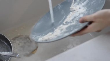 Üzerinde un ve hamur kalıntısı olan gri bir tabak beyaz bir lavaboda akan suyun altında yıkanır. Mutfak temizleme sahnesinde musluk duş moduna geçer.