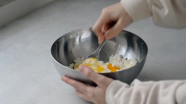 Bir kadın, parlak sarı bir yumurtayı kaba süzme peynirle metal bir kasede karıştırıyor, şurubu hazırlıyor, süzme peynirli krep. Geleneksel bir Slav tatlı tarifi hazırlama.