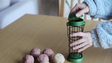 Bir kadın silindirik bir kuş yemliğini evdeki ahşap bir masada asılı duran şişman toplarla dolduruyor ve kışın kuşlara yiyecek sağlamak için onu hazırlıyor..
