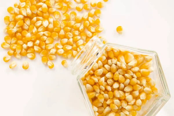 Sarı patlamış mısır taneleri beyaz arka planlı altıgen cam kavanozun yanına saçılmış.