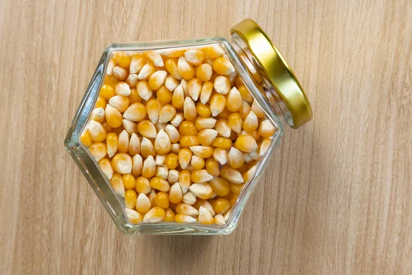 Altıgen cam bir kavanozda sarı kurutulmuş mısır taneleri açık renkli ahşap bir masada altın kapaklı.