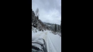Kışın karlı bir dağ yolunda giden bir aracı gösteren video. Orman yamaçları, taze kar ve bulutlu gökyüzü uzak bir kış yolculuğu sırasında sessiz ve soğuk bir atmosfer yaratır..