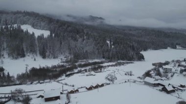 Karla kaplı orman ve tepelerle çevrili sessiz bir dağ köyünün hava manzarası. İnsansız hava aracı görüntüleri kırsal evleri, donmuş manzarayı ve sakin mevsimsel atmosferi gösteriyor..