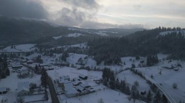 Drone 'un hava görüntüleri karlı köyün üzerinde ve gün batımında dolambaçlı yolda uçuyor. Soğuk dağ vadisinde evler, ağaçlar ve yumuşak akşam ışıklarıyla kışın kırsal alan.