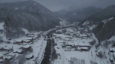 Dar bir kış vadisinde, soğuk mevsimsel hava koşullarında nehir, ev ve orman manzaralı bir dağ köyünün üzerinde uçan insansız hava aracı görüntüsü..